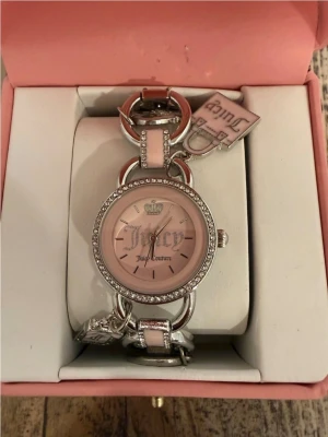 Juicy Couture rosa klocka med strass - Juicy Couture klocka med rosa urtavla och silverfärgade detaljer. Runt urtavlan sitter gnistrande strass och armbandet är i ljusrosa och silver. Klockan har dekorativa berlocker och levereras i original ask. Perfekt accessoar för dig som gillar glam.