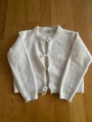 Beige kofta med knytning framtill - Mysig beige kofta från SAMSA med rund halsringning och tre knytband framtill. Koftan är stickad i ett mjukt material och har långa ärmar samt ribbade muddar vid ärmslut och hals. Perfekt för lager på lager och en avslappnad look.