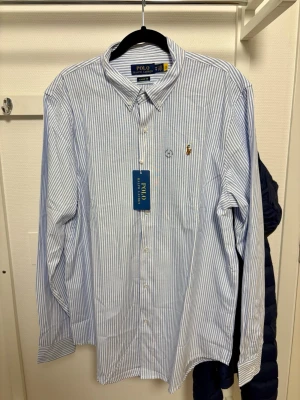 Blårandig skjorta från Polo Ralph Lauren - Klassisk blårandig skjorta från Polo Ralph Lauren i bomull. Helt ny och oanvänd, endast testad. Classic Fit M gör att den är lite rymligare än Slim Fit och passar bättre för en mer relaxed stil. Lika lång som en vanlig Slim Fit M (ganska lång så passar en som har L också)