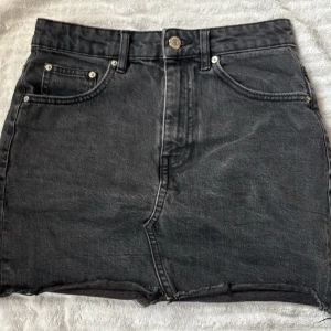Svart jeanskjol med rå kant - Säljer en svart jeanskjol med fem fickor och rå, fransig kant nedtill. Kjolen har klassisk knapp- och dragkedjestängning samt bälteshällor. Perfekt till en avslappnad och trendig look.
