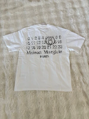 Maison Margiela vit t-shirt  - Vit t-shirt från Maison Margiela med svart grafiskt tryck och logga på ryggen. Klassisk passform med korta ärmar och rund halsringning. Tillverkad i mjuk bomull som känns skön mot huden. Perfekt för dig som gillar minimalistisk och stilren design.