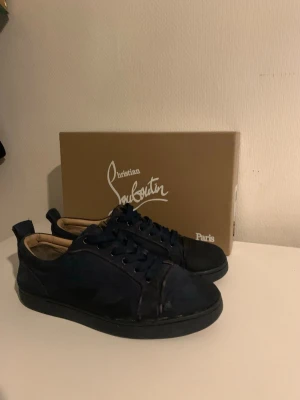 Mörkblå Christian Louboutin sneakers - Snygga mörkblå sneakers från Christian Louboutin med klassisk låg profil och ikoniska röda sulor. Skorna har svarta snören och detaljer i mocka, vilket ger en exklusiv känsla. Perfekta för dig som vill sticka ut med stilrena och lyxiga sneakers.