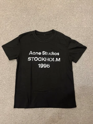 Svart Acne Studios t-shirt - Säljer en svart t-shirt från Acne Studios med vit text på bröstet där det står 'Acne Studios STOCKHOLM 1996'. Klassisk rund hals och korta ärmar. Perfekt för dig som gillar stilrena och trendiga plagg.