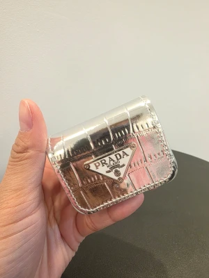 Silverfärgad AirPods-skal från PD - Snyggt AirPods-skal från PD i metallic silver. Fodralet har en trekantig metall-logga framtill och stilren design. Perfekt för och tillverkad i glansigt material som sticker ut. 