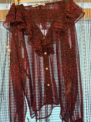 Röd- och svartmönstrad blus med volang - Snygg transparent blus från Zara i rött och svart med djurmönster. Blusen har volangdetaljer längs v-ringningen och knappar framtill. Korta ärmar och lätt, luftig känsla. Perfekt för dig som gillar att sticka ut med din stil.