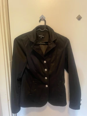 Svart ridkavaj från Equipage - Stilren svart ridkavaj från Equipage med klassisk krage och fyra silverfärgade knappar framtill. Kavajen har två fickor och är figursydd för en snygg look.