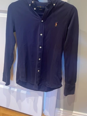 Mörkblå skjorta från Ralph Lauren - Klassisk mörkblå skjorta från Ralph Lauren med knappslå och vita knappar. Skjortan har lång ärm, button-down krage och den ikoniska broderade loggan på bröstet. Materialet är knit oxford som ger en mjuk och bekväm känsla.