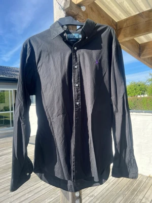 Svart skjorta från Polo Ralph Lauren - Klassisk svart skjorta från Polo Ralph Lauren med lila broderad logga på bröstet. Skjortan har knappar framtill och lång ärm. Perfekt till jeans eller chinos för en clean look.