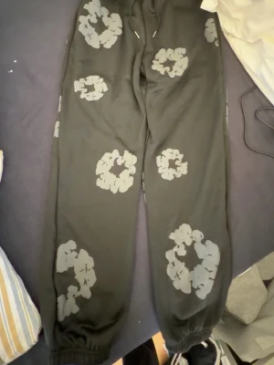 denim tears byxor - Säljer ett par svarta mjukisbyxor med stora gråa blommönster över hela byxorna. Byxorna har resår i midjan och vid bensluten samt dragsko i midjan för extra komfort. Perfekta för en chill och avslappnad stil.