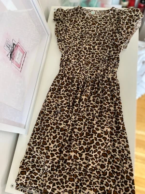 Leopardmönstrad klänning med volang i ärmarna - Snygg leopardmönstrad klänning med korta ärmar och markerad midja. Klänningen har en beige bas med bruna och svarta fläckar och är tillverkad i ett tunt, mjukt tyg med innerfoder. Perfekt för dig som gillar djurmönster och vill sticka ut.