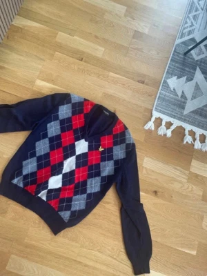 Mörkblå vintage argyle stickad tröja från lyle scott - Snygg mörkblå stickad tröja med klassiskt argylemönster i rött, grått och vitt på framsidan. Tröjan har långa ärmar, ribbade muddar och en liten gul broderad detalj på bröstet. 