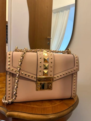 Ljusrosa handväska med nitar Michael Kors - Säljer en ljusrosa handväska från Michael Kors med guldfärgade nitar och kedjerem. Väskan har ett elegant lås framtill och detaljer i guld. Tillverkad i slätt läder och har en stilren, boxig form som ger en lyxig känsla.