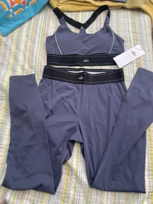 Grått träningsset från Alo - Snyggt träningsset från Alo i grått med svarta och vita detaljer. Setet består av en sport-bh med korsade axelband och matchande tights med bred resår i midjan. Materialet är stretchigt och följsamt, perfekt för gymmet eller yoga.