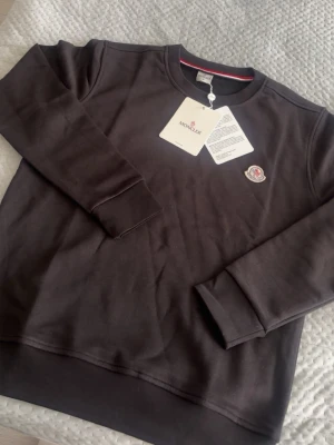 Svart sweatshirt från Moncler - Snygg svart sweatshirt från Moncler med rund halsringning och Moncler-logga broderad på bröstet. Tröjan har långa ärmar och ribbade muddar vid ärmslut och nederkant. Tillverkad i mjukt bomullsmaterial för en skön känsla.