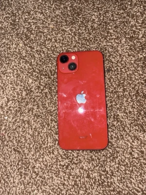 IPhone 13 - Snygg röd iPhone med två kameror på baksidan och klassisk Apple-logga i silver. Telefonen har en stilren design med rundade hörn och baksida i glas. Perfekt för dig som gillar färg och vill ha en modern smartphone.