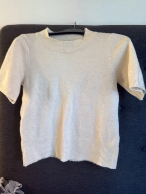 Beige stickad t-shirt - En enkel och stilren beige stickad t-shirt med korta ärmar och rund hals. Tröjan har ribbstickade muddar vid hals, ärmslut och nederkant. Perfekt för lager på lager och passar till det mesta.