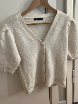 Vit kortärmad kofta från Gina Tricot - Söt vit kofta från Gina Tricot med korta puffärmar och v-ringning. Koftan har ribbade muddar, knappar framtill och är stickad i ett mjukt material. Perfekt att slänga över en topp för en chill look. Typ helt oanvänd!