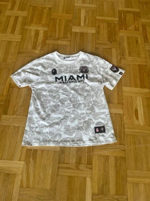 Vit camo Miami BAPE t-shirt - Säljer en vit t-shirt från BAPE med Miami-tryck framtill och stor svart 93:a samt BAPE-tryck på ryggen. T-shirten har ett grått camo-mönster över hela plagget och flera patchar på bröstet och ärmen. Klassisk rund hals och korta ärmar.
