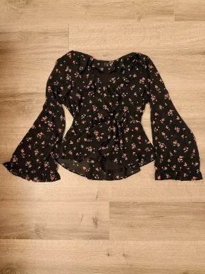 Blommig blus från H&M Divided S - Svart blus med små rosa blommor från H&M Divided. Blusen har långa, utsvängda ärmar och volangdetaljer framtill. Tillverkad i ett lätt och luftigt tyg som ger en snygg siluett. 