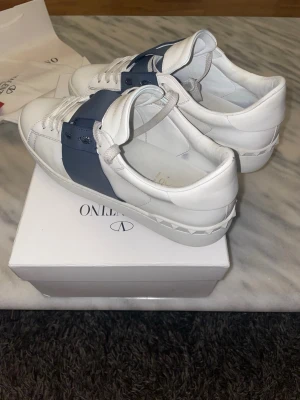 Valentino - Snygga vita sneakers med bred blå rem över snörningen och coola nitar runt sulan. Skorna har en klassisk låg profil och är gjorda i slätt läder med vita skosnören. Perfekta för dig som vill ha en stilren men ändå unik look. Priset är inte hugget i sten!!