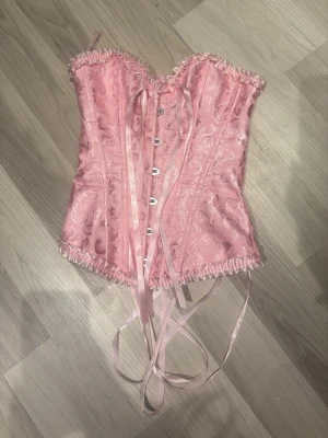 Rosa korset 💕 - Säljer en helt fantastisk och oanvänd rosa korsett. Tyget har ett vackert, vävt brokad/paisleymönster med fin lyster  vilket ger ett exklusivt och elegant intryck. Korsetten pryds även av söta volangdetaljer i satin längs över- och nederkanten. Den har klassisk korsettsnörning med rosa satinband i ryggenä, vilket gör att passformen kan justeras precis som man vill ha den. Framtill 