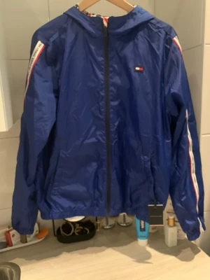 Blå vindjacka från Tommy Hilfiger - Snygg blå vindjacka från Tommy Hilfiger med huva och dragkedja. Jackan har vita och röda detaljer längs ärmarna och ett stort Hilfiger-tryck på insidan. Liten logga på bröstet och två fickor framtill. Perfekt för blåsiga dagar och streetwear-stil.
