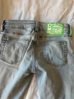 Cheap monday jeans - Storlek 27/32.  Säljer ett par ljusblå jeans med utsvängda ben och klassisk femficksmodell. Jeansen har en unik grön patch med tryck bak i midjan och slitningar framtill för en cool look. Perfekta för dig som gillar streetwear och vill sticka ut lite extra.