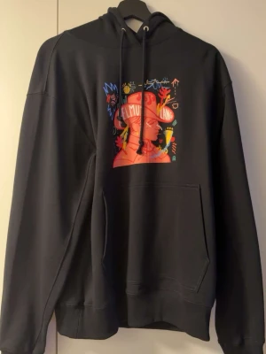 Svart hoodie från Helmut Lang  - Cool svart hoodie med stor färgglad grafisk illustration på bröstet som föreställer ett ansikte och olika symboler. Hoodien har huva med dragsko och en stor magficka framtill. Perfekt för dig som gillar streetwear och unika prints.
