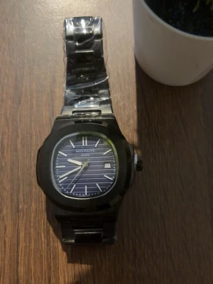 Klocka  - Stilren klocka med svart metallarmband och blå urtavla med vita detaljer. Urtavlan har datumvisning och tydliga markeringar. Klassisk design som passar till många stilar.