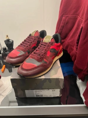 Valentino rockeunners röd camo - Snygga sneakers från Valentino i röd och vinröd camo-design med svarta och grå detaljer. Skorna har lågt skaft, svart insida och beige sula. Materialmix av mocka, läder och textil ger en exklusiv känsla och cool look. Pris kan diskuteras 