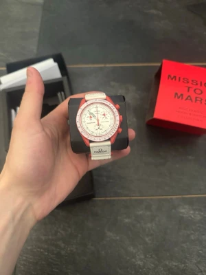 Omega x Swatch Mission to Mars klocka - Säljer en Omega x Swatch Mission to Mars klocka med vit urtavla, röda detaljer och vit textilrem. Klockan har kronograf och tydliga markeringar, samt coola röda visare. Perfekt för dig som gillar unik design och vill sticka ut.