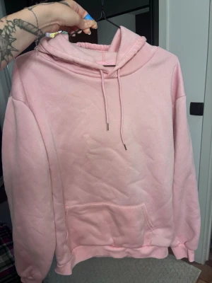 Ljusrosa hoodie med huva och ficka - Mysig ljusrosa hoodie med stor känguruficka framtill och justerbar dragsko i huvan. Tillverkad i mjukt material och har ribbade muddar vid ärmar och nederkant. Lite nopprig se bild 