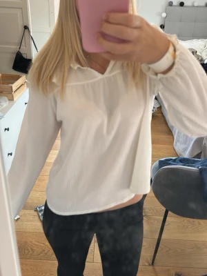 Vit blus med volangkrage - Söt vit blus med lång ärm och volangdetaljer vid kragen. Blusen har en lös passform och är tillverkad i ett lätt, strukturerat tyg som ger en luftig känsla. Perfekt att styla med jeans eller kjol för en trendig look.
