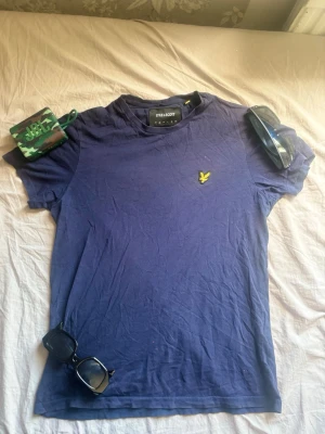 Blå t-shirt från Lyle & Scott - Snygg blå t-shirt från Lyle & Scott med klassisk gul broderad logga på bröstet. T-shirten har rund hals och korta ärmar, perfekt för en avslappnad stil. Tillverkad i mjuk bomull för skön komfort.Pris kan diskuteras.