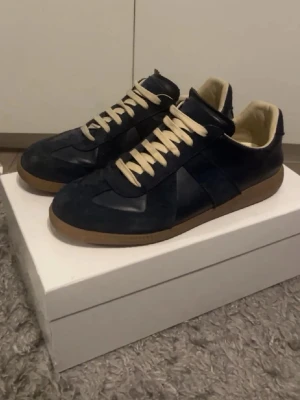 Maison margiela - Snygga mörkblå sneakers med beige snören och kontrasterande gummisula. Ovandelen är i mocka och läder, vilket ger en schysst mix av material. Låg modell som passar till det mesta och har en klassisk siluett.