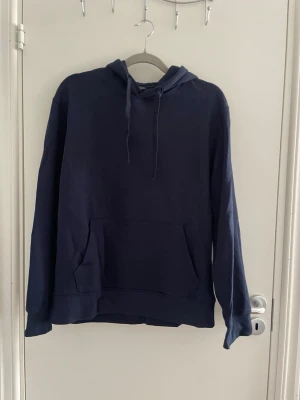 Mörkblå hoodie med ficka - En enkel och stilren mörkblå hoodie med huva och dragsnören. Hoodien har en stor magficka framtill och ribbade muddar vid ärmslut och nederkant. Perfekt för en avslappnad look och passar till det mesta. Materialet känns mjukt och bekvämt.