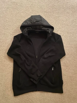 Cardigan  - Säljer en svart cardigan med dragkedja och huva i glansigt tyg. Cardiganen är stickad och har två fickor med dragkedja framtill. Perfekt för en chill och stilren look.