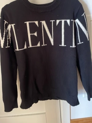 Svart stickad tröja från Valentino - Snygg svart stickad tröja från Valentino med stort vitt logotyp-tryck över bröstet. Tröjan har rund halsringning, långa ärmar och ribbade muddar vid ärmslut och nederkant. Tillverkad i Italien och perfekt för dig som gillar exklusiv streetstyle.