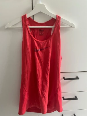 Röd Nike Dri-FIT träningstopp - Snygg röd träningstopp från Nike med Dri-FIT-material som andas. Linnet har en klassisk racerback-design och ett tryckt Nike Swoosh på bröstet. Perfekt för gymmet eller löprundan när du vill ha något lätt och bekvämt.