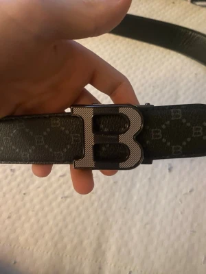 Svart Balmain bälte med B-spänne - Snyggt svart bälte från Balmain med stort B-format metallspänne. Bältet har diskret mönster med små B-loggor över hela remmen och är gjort i skinn. Perfekt accessoar för att lyfta din outfit.