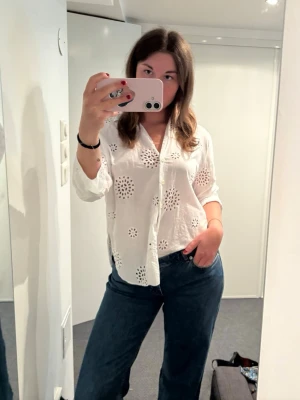 Vit skjorta med broderade hålmönster - Säljer en vit skjorta med unika broderade hålmönster och knappar framtill. Skjortan har lång ärm och en avslappnad passform, perfekt för en chill och stilren look. Tillverkad i lätt bomull som känns skön mot huden.