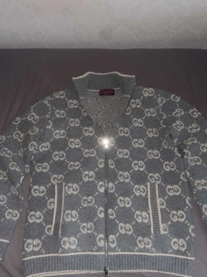 Grå stickad ziptröja från Gucci - Säljer en grå stickad ziptröja från Gucci med hel dragkedja och hög krage. Tröjan har ett mönster med vita GG-loggor över hela plagget samt ribbade muddar och fickor framtill. Perfekt för dig som gillar exklusiv streetwear och snygga detaljer.