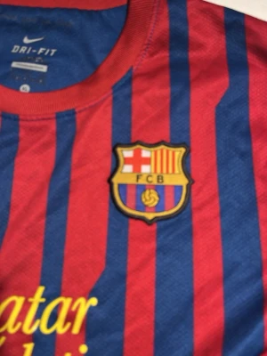 Barcelona fotbollströja Nike röd/blå - Säljer en klassisk FC Barcelona matchtröja från Nike med Qatar Foundation-tryck. Tröjan är röd och blå med smala vertikala ränder, klubbmärke på bröstet och korta ärmar. Tillverkad i lätt och ventilerande polyester, perfekt för fotboll eller samling.