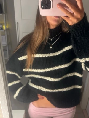 Svart stickad tröja med vita ränder - Mysig svart stickad tröja med breda vita ränder på både kropp och ärmar. Tröjan har hög hals och en avslappnad passform. Perfekt för kalla dagar och enkel att styla till olika outfits.