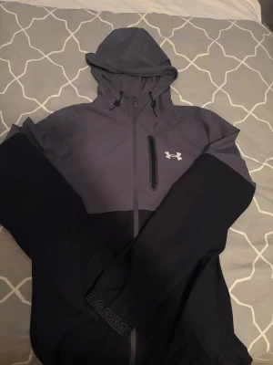 Sällsynt under armour jacka - Snygg tvåfärgad vindjacka från Under Armour i mörksvart och grått. Jackan har huva med dragsko, hel dragkedja framtill och en bröstficka med dragkedja. Tillverkad i lätt och tåligt material, perfekt för blåsiga dagar. Under Armour-logga på bröstet.