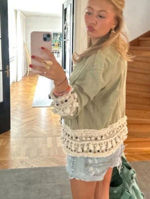 Grön jacka med spets och tofsar - Snygg ljusgrön jacka med dekorativa spetsdetaljer och tofsar längs ärmslut och nederkant. Jackan har två bröstfickor och en avslappnad passform. Perfekt för dig som vill sticka ut med en unik och trendig look.