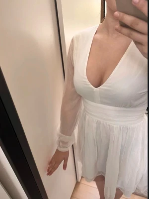 Vit kort omlottklänning med v-ringning - Säljer en vit kort klänning med v-ringning och långa, transparenta ärmar. Klänningen har omlott-look i midjan och är gjord i ett lätt, flödande material som ger en luftig känsla. Perfekt för sommarens alla tillfällen!