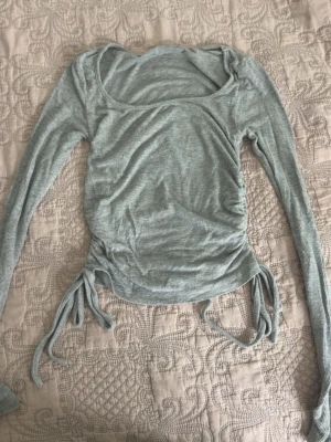 Ljusgrå ribbad topp med snörning - Trendig ljusgrå ribbad topp med långa ärmar och snygg snörning i sidorna. Toppen har en något croppad passform och en fyrkantig ringning. Perfekt för att lyfta din vardagsstil med en modern touch.