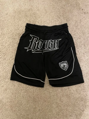 Svarta Rough shorts med vit piping - Svarta shorts från Rough med vit piping längs kanterna och stort tryck framtill. Elastisk midja med snörning och en patch med logga på ena benet. Perfekta för en sportig och avslappnad stil.