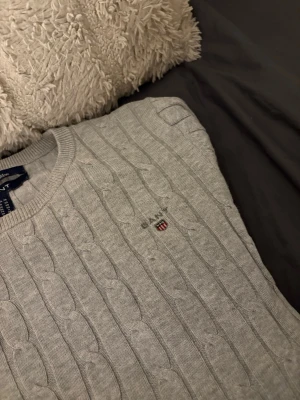 Ljusgrå kabelstickad tröja från Gant - Snygg ljusgrå kabelstickad tröja från Gant med rund halsringning och diskret logga broderad på bröstet. Tröjan har långa ärmar och ribbade muddar vid ärmslut och nederkant. Perfekt för lager på lager och klassisk stil.